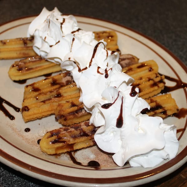 El Nopal's churros dessert, topped with whipped cream and chocolate syrup, on a white plate.