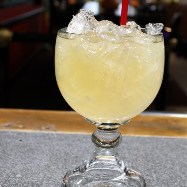 A large glass of El Nopal's Mexican margarita, filled with ice and a red straw, on a bar.