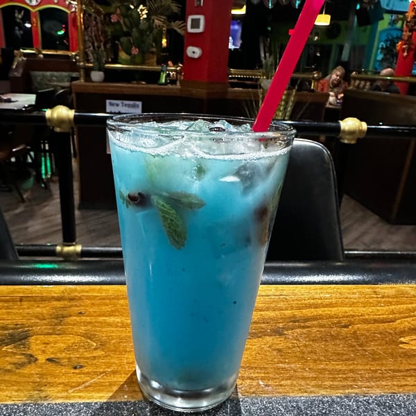 A vibrant blue cocktail with ice, mint, and a red straw sits on a wooden bar.